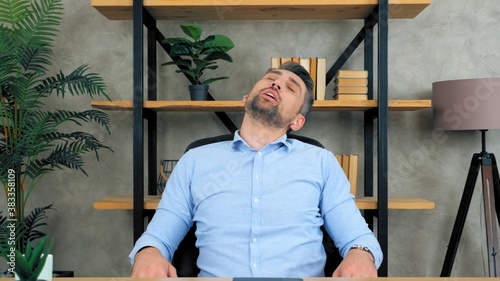 Tired gray-haired bearded business man manager SEO freelancer wears wireless earphones asleep on chair at table after long hard work at home office