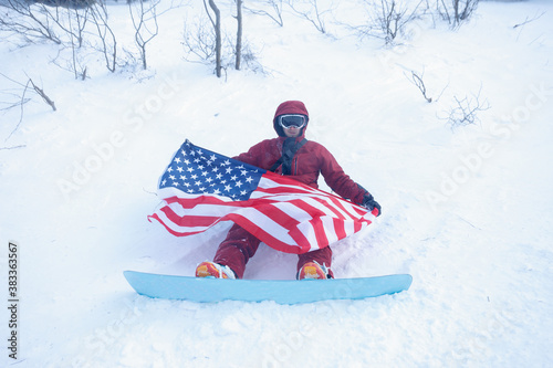 winter, leisure, sport and people concept - snowboarder sits high in the mountains on the edge of the slope and looks into the distance. snowboarder holding american flag