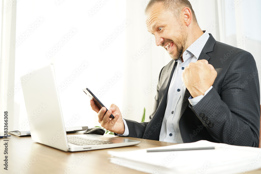 a businessman man in the office has read a news or message in his smartphone and brightly expresses emotions of joy, surprise and happiness with a smile a businessman man in the office has read a news or message in his smartphone and brightly expresses emotions of joy, surprise and happiness with a smile