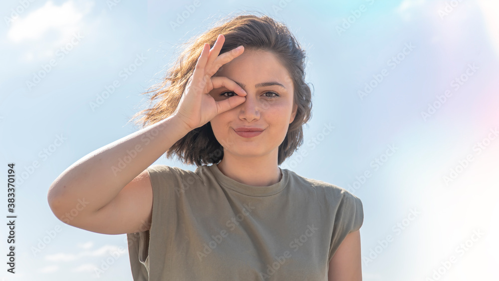 Charming young woman doing ok gesture with her hand and looking through OK sign smiling with happy face against sky background. Facial expression.