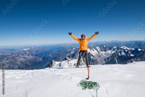 Happy smiling rope team man with climbing axe dressed orange mountaineering clothes,boots with crampons and safety harness rised a hands on the Mont Blanc (Monte Bianco) summit 4,808m