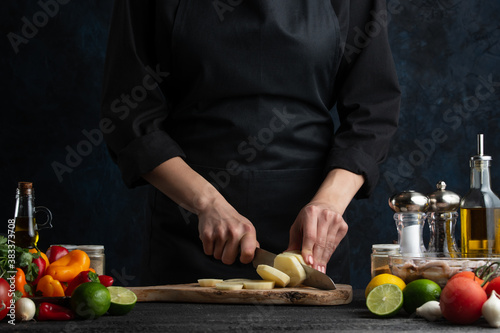 The chef in black uniform cuts with knife raw potato for preparing dinner on dark blue background. Backstage of cooking meal at commercial kitchen. Concept of eco food.