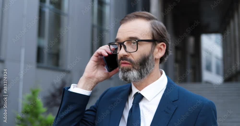 Caucasian middle-aged man in glasses talking on mobile phone and drinking coffe to-go in morning outdoors. Close up of businessman speaking on cellphone and sipping hot drink at business center. Caucasian middle-aged man in glasses talking on mobile phone and drinking coffe to-go in morning outdoors. Close up of businessman speaking on cellphone and sipping hot drink at business center.