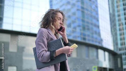 Side view of young business woman eating on the go and talking on mobile phone against background of office building