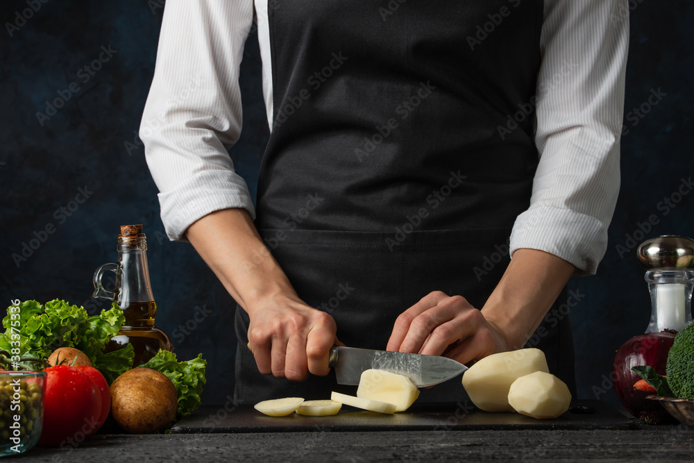 The professional chef in black apron cuts with knife potato on black chopped board on dark blue background. Backstage of preparing restaurant meal for dinner. Food concept. Close-up. The professional chef in black apron cuts with knife potato on black chopped board on dark blue background. Backstage of preparing restaurant meal for dinner. Food concept. Close-up.