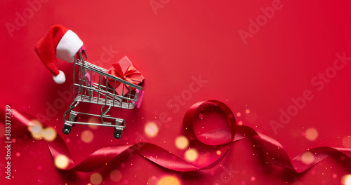 on a red background lies a cart from a store on wheels with a red gift inside and a hat of Santa Claus on the handle
