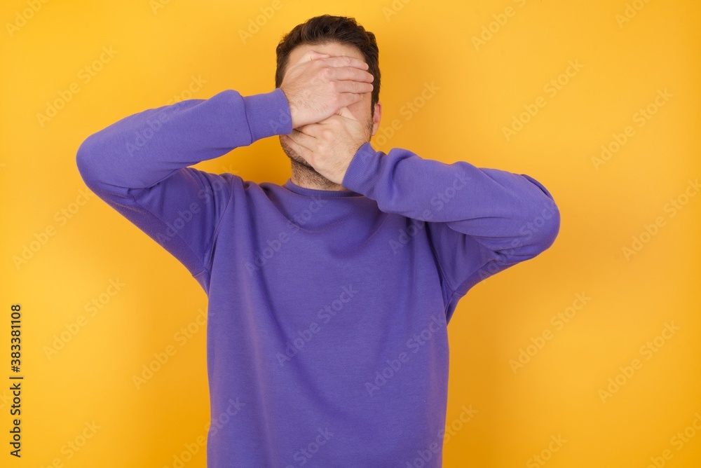 Handsome man with sweatshirt over isolated yellow background Covering eyes and mouth with hands, surprised and shocked. Hiding emotions. Handsome man with sweatshirt over isolated yellow background Covering eyes and mouth with hands, surprised and shocked. Hiding emotions.