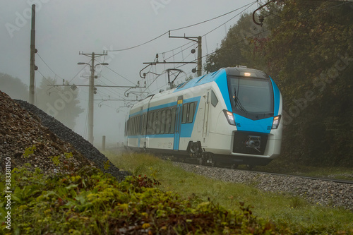 Modern and new passenger commuter Train in white and blue color is traveling on a single track railway line between the green leaves on a cold foggy day.