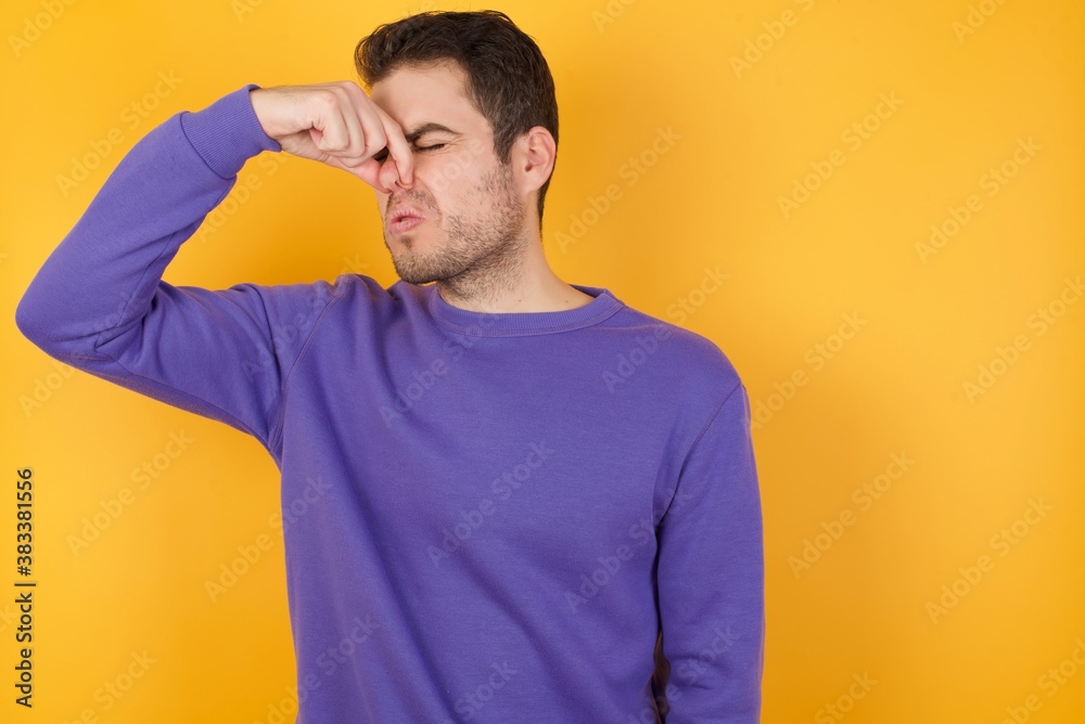 Handsome man with sweatshirt over isolated yellow background smelling something stinky and disgusting, intolerable smell, holding breath with fingers on nose. Bad smell Handsome man with sweatshirt over isolated yellow background smelling something stinky and disgusting, intolerable smell, holding breath with fingers on nose. Bad smell