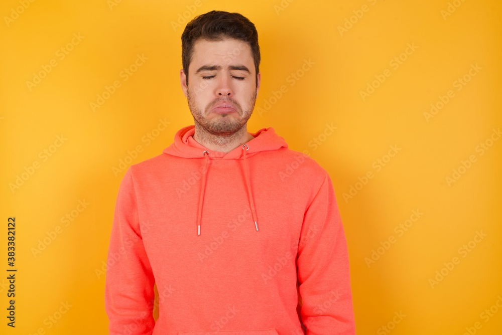 Dismal gloomy rejected Handsome man with sweatshirt over isolated yellow background has problems and difficulties, curves lower lip and closes eyes in despair, being in depression Dismal gloomy rejected Handsome man with sweatshirt over isolated yellow background has problems and difficulties, curves lower lip and closes eyes in despair, being in depression