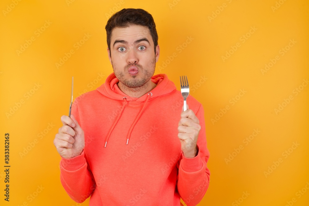 Handsome man with sweatshirt over isolated yellow background hungry girl holding in hand fork knife want tasty yummy pizza pie Handsome man with sweatshirt over isolated yellow background hungry girl holding in hand fork knife want tasty yummy pizza pie