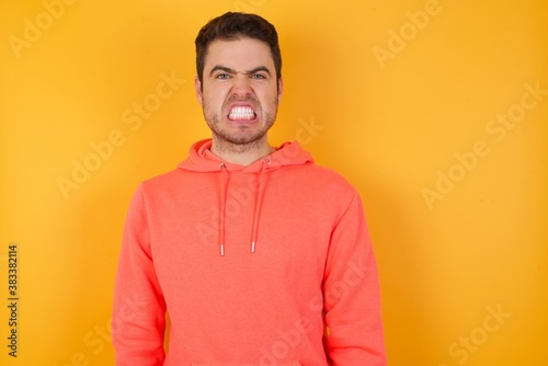 Handsome man with sweatshirt over isolated yellow background keeps teeth clenched, frowns face in dissatisfaction, irritated because of much duties. © Roquillo