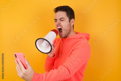 Crazy impressed Handsome man with sweatshirt over isolated yellow background use cell phone reads incredible news on social media information stare, screams wow omg wear stylish outfit