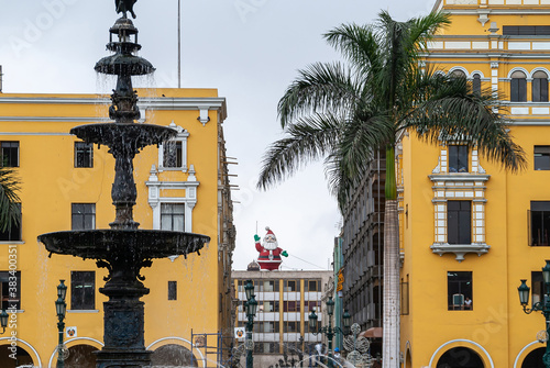 Lima, Peru - December 4, 2008: Red Santa Claus mascotte on roof visible through Pje De Jose Olaya alley between 2 yellow buildings on Jiron Huallaga blvd under light blue sky. Green foliage and founta photo