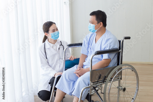 Asian woman doctor asking about health with a man patient who sit on wheel chair in hospital. They wear medical face mask to protect respiration system. (Covid-19)