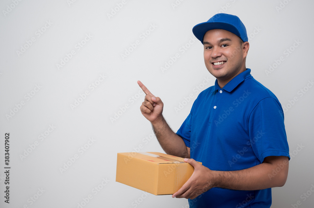 Young smiling asian delivery man in blue uniform holding box parcel cardboard and pointing finger on blank space isolated white background.