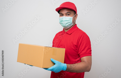 Young asian delivery man in red uniform wearing protection mask and medical rubber gloves giving parcel cardboard to customer on isolated white background. Safety deliver concept.