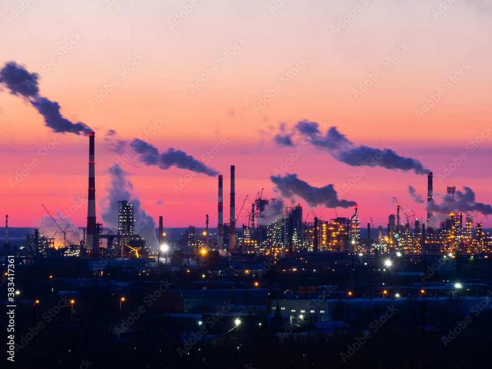 Refinery at sunset. Pipes of a thermal power station and an oil refinery, illuminated plant installations on a red sunset sky. Blue smoke from pipes of an oil refinery and plastic