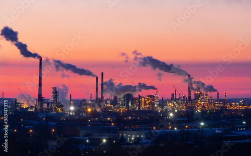 Refinery at sunset. Pipes of a thermal power station and an oil refinery, illuminated plant installations on a red sunset sky. Blue smoke from pipes of an oil refinery and plastic