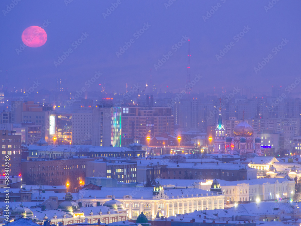 Fototapeta premium Big red full moon over Omsk, full moon over the city center. Lenin street, Assumption Cathedral and a smoking oil factory against the backdrop of the city with the setting moon in the morning.