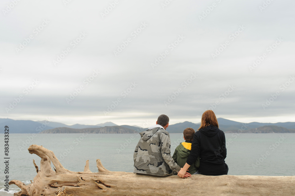 family of three sits back on an old tree without bark on sandy beach and admires the blue clear sea and mountains, family vacations, domestic tourism
