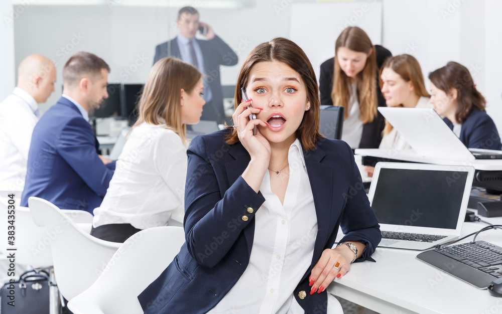 Obraz premium Smiling young female emotionally talking on mobile in busy modern open plan office