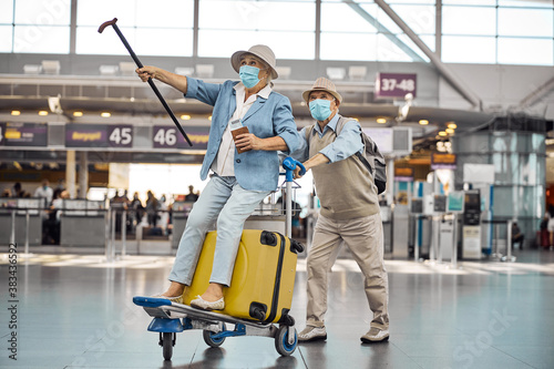 Male pensioner wheeling his wife on a baggage trolley