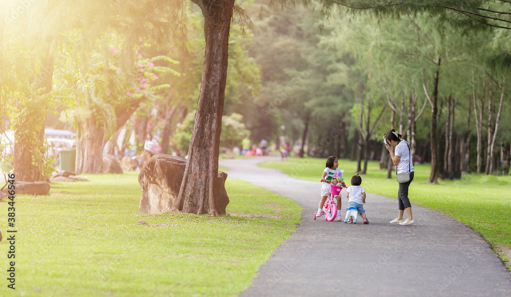 Fototapeta premium Family in park concept. Mother and her daughter play at park together. Mother shot her daugther with camera. Little girls relax with bicycle on road in park. Young mother shot picture her daughter