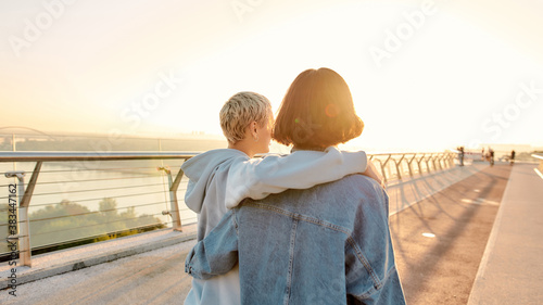 Lesbian couple standing on the bridge, embracing each other while admiring the sunrise together. Homosexuality, LGBT and love concept