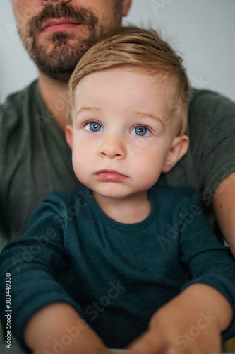 Youngster sitting in father s lap and looking at something with his beautiful big blue eyes.