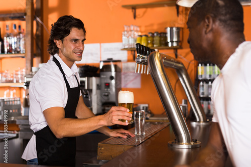 Young man barman giving beer with foam to man client in bar
