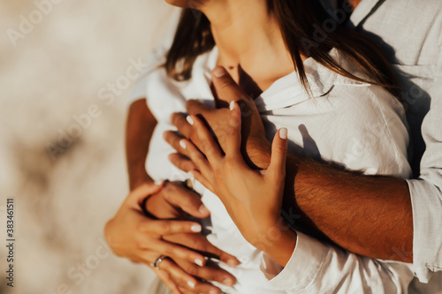 Cropped photo of young guy and girl in white clothes. Guy gently hug his girlfriend. Hands - close-up.