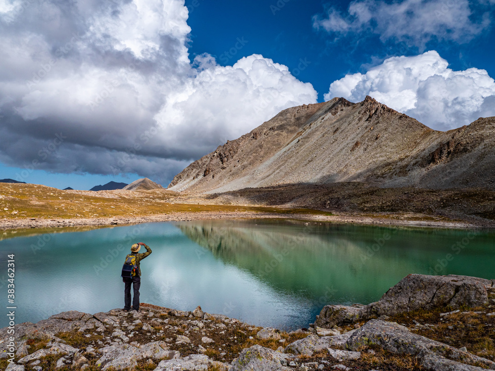 Male tourist near the shore of a high mountain lake. Large white clouds and mountains are reflected in the turquoise water. Kazakhstan, Kensu gorge Male tourist near the shore of a high mountain lake. Large white clouds and mountains are reflected in the turquoise water. Kazakhstan, Kensu gorge