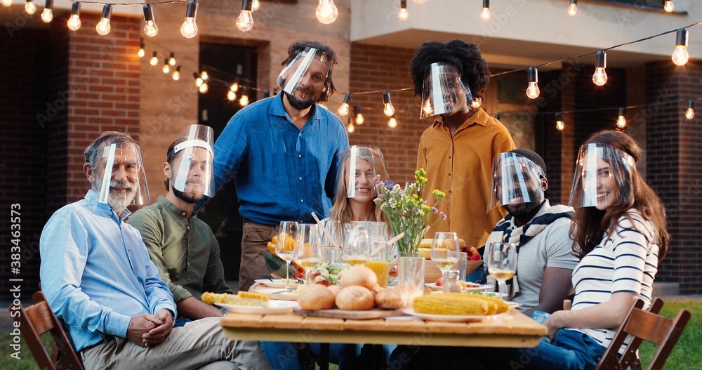 Portrait of mixed-races people in face shields at dinner table in back yard and smiling to camera. Multi ethnic family at picnic in court outdoors. Pandemic communication of generations. Portrait of mixed-races people in face shields at dinner table in back yard and smiling to camera. Multi ethnic family at picnic in court outdoors. Pandemic communication of generations.