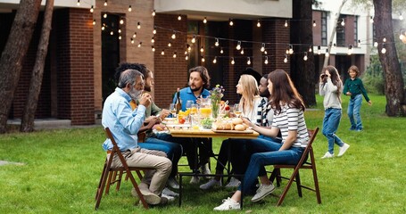 Happy Caucasian family sitting at table with meal outdoor at picnic and talking. Joyful young and old people having dinner and having nice communication. Weekend celebration gathering.