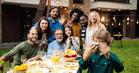 Mixed-races happy family at party dinner outdoor in yard smiling and posing to smartphone camera while small boy taking selfie photo. Multi ethnic people making photos together at barbeque Celebration