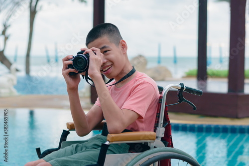 Asian special child on wheelchair is holding camera take photo at swimming pool in front of sea beach,Boy happiness in holidays with family time on the travel, Lifestyle of happy disabled kid concept.