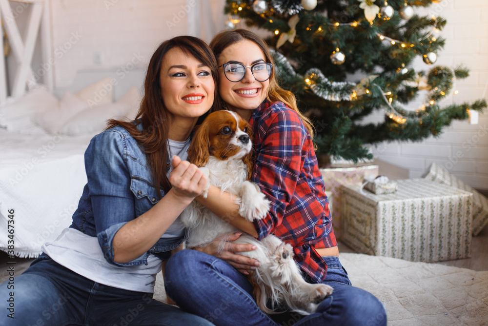 two girlfriends with dog are sitting on the floor smiling in a bright room on the background of a Christmas tree