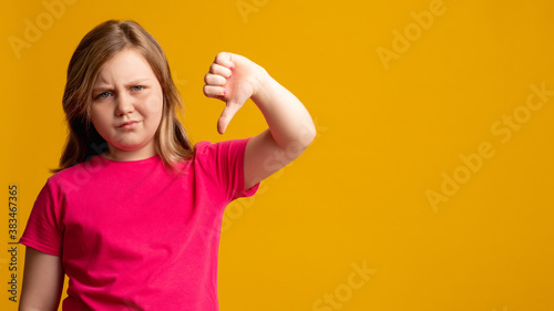 Dislike gesture. Disappointed child. Wrong choice. No answer. Dissatisfied young girl in pink criticizing idea with thumb down looking at camera isolated on orange copy space background.