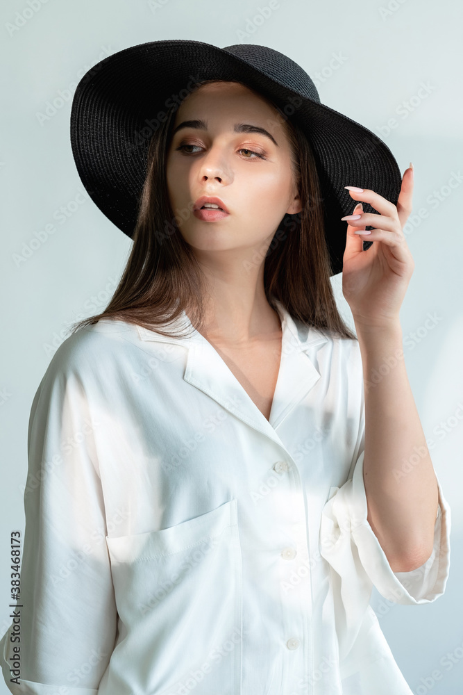 Fashion look. Model posing. Beautiful young woman in white shirt touching black wide-brimmed hat in sun light glow isolated on neutral. Season trend. Female elegance. Fashion look. Model posing. Beautiful young woman in white shirt touching black wide-brimmed hat in sun light glow isolated on neutral. Season trend. Female elegance.