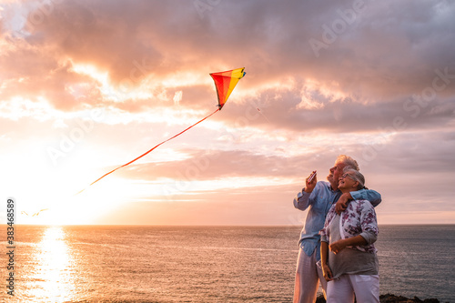 two happy and cheerful senior having fun enjoying and playing with a flying kite at the beach with the sunset and the sea at the background - active lifestyle people outdoors photo