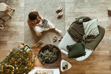 Young beautiful woman sitting on carpet and work on laptop in cozy comfortable living room decorated for Christmas / New Year holidays celebration. Christmas tree, sofa, blanket, decorations. Top view