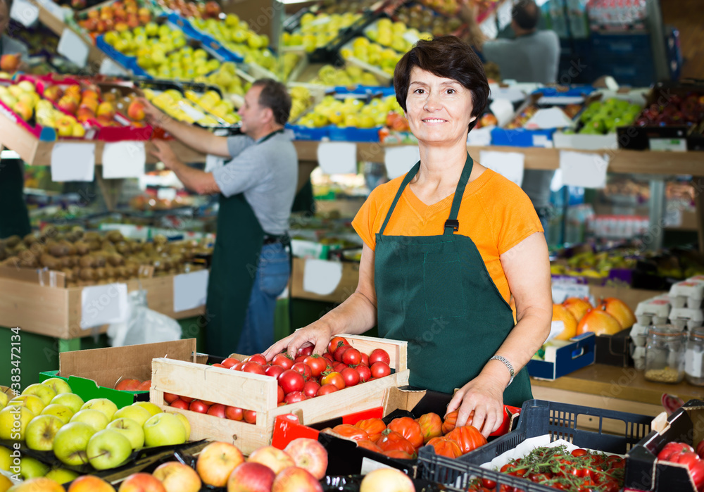 Obraz premium Positive female seller is standing with box with tomatos in the supermarket