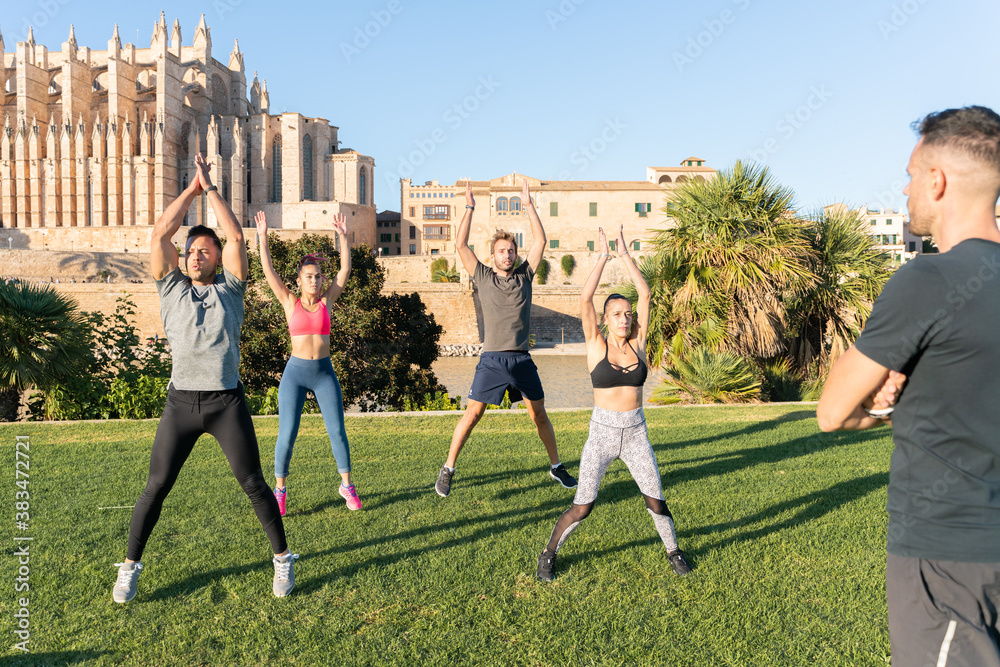 Group of young people practicing jumping jack exercise in fitness session, instructed by their personal trainer in the park in front of Palma Cathedral