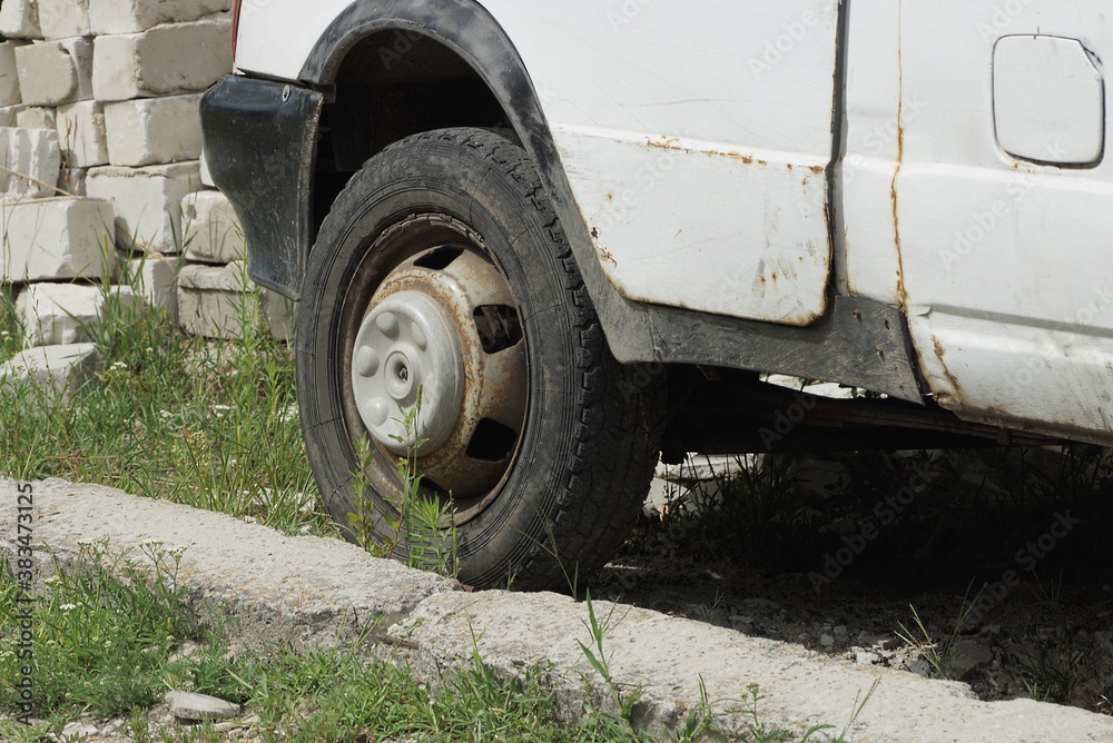 part of a white minibus with one black dirty wheel   stands on the ground and green grass on the street
