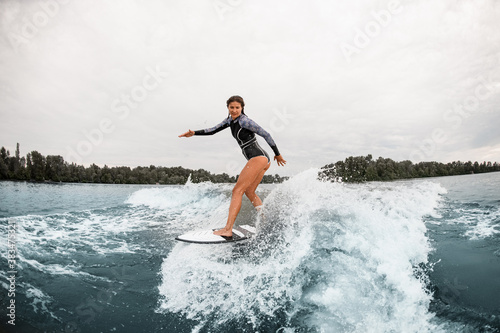 attractive woman in black wetsuit stand on a surfboard and rides on wave
