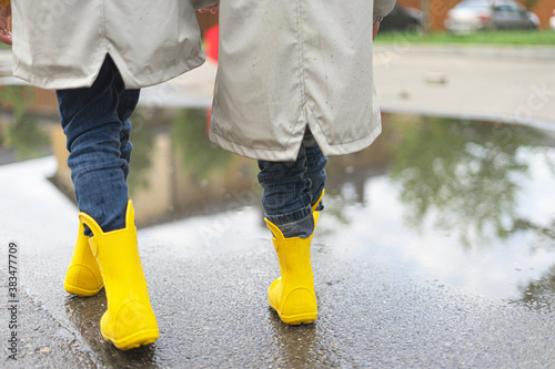 Children's feet in yellow rubber rain boots go down a puddle in the street