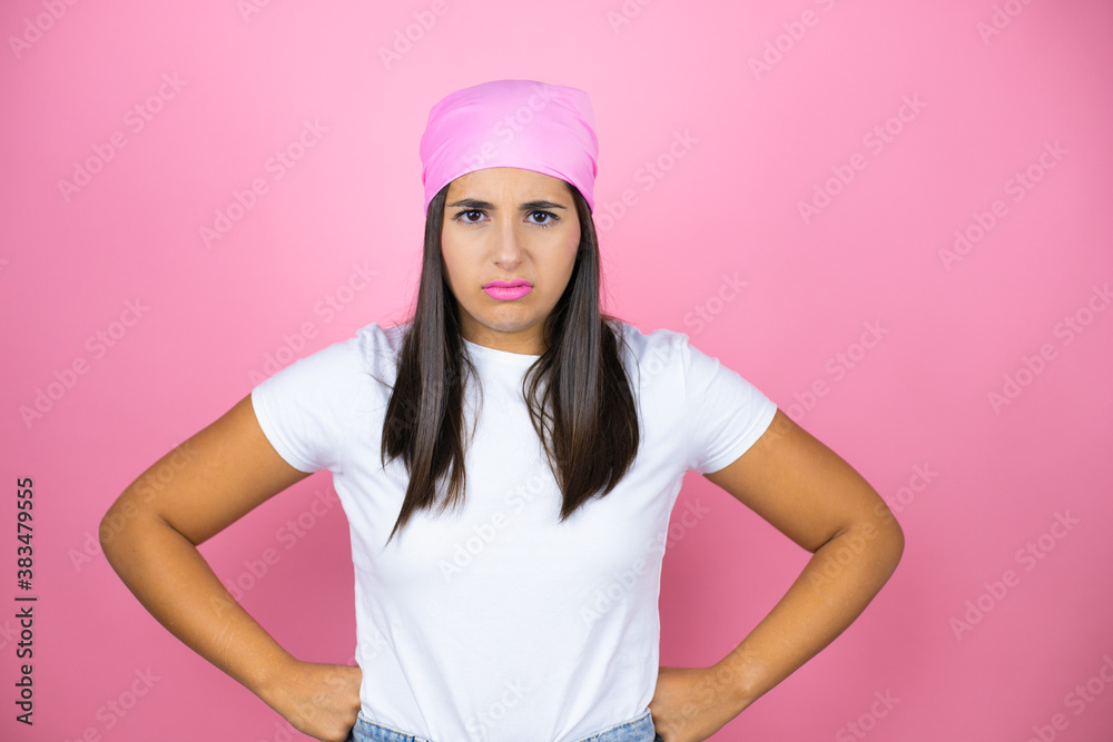 Young beautiful woman wearing pink headscarf over isolated pink background skeptic and nervous, disapproving expression on face with arms in waist