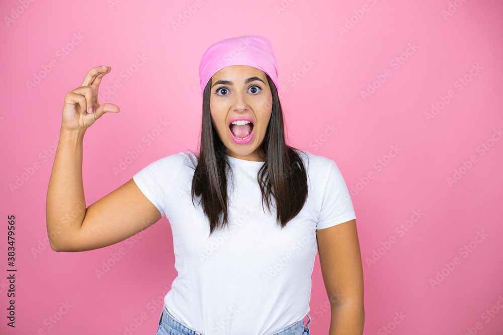 Young beautiful woman wearing pink headscarf over isolated pink background smiling and confident gesturing with hand doing small size sign with fingers looking and the camera. Measure concept. Young beautiful woman wearing pink headscarf over isolated pink background smiling and confident gesturing with hand doing small size sign with fingers looking and the camera. Measure concept.