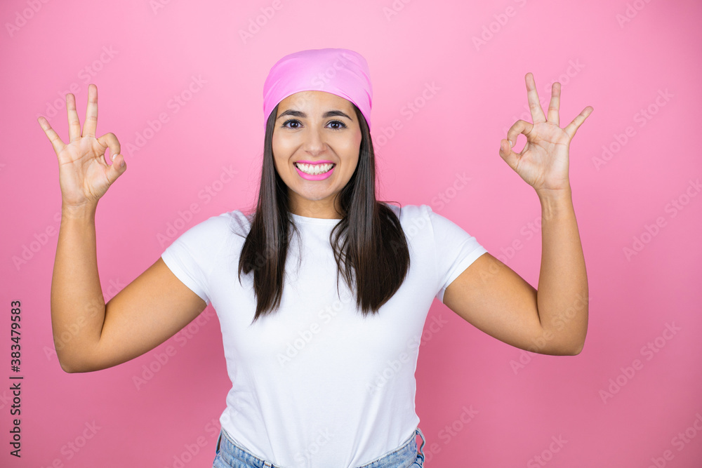 Young beautiful woman wearing pink headscarf over isolated pink background doing ok sign with fingers and smiling, excellent symbol Young beautiful woman wearing pink headscarf over isolated pink background doing ok sign with fingers and smiling, excellent symbol